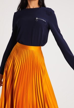 Kobieta w granatowej bluzce z długim rękawem, z zamkiem przy kieszeni na piersi i wysokim stanem, plisowaną, jaskrawo pomarańczową spódnicą.