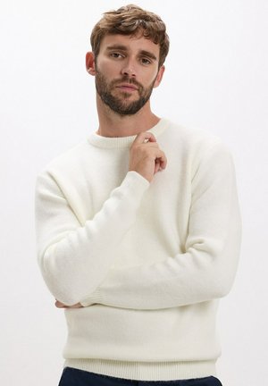 Pull en tricot blanc avec un col rond et des manches longues. Texture douce et poignets côtelés. Porté sur un pantalon noir, avec une coupe uniforme.