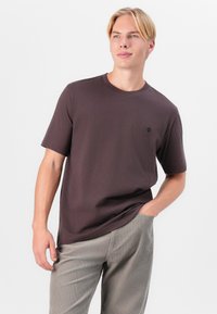Dark brown cotton t-shirt with short sleeves, crew neck, and a small logo detail on the chest. Worn with light grey corduroy pants.