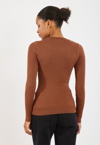 Bronzas svītrains džemperis ar garām piedurknēm, apaļu kakla izgriezumu un pieguļošu siluetu. Kombinēts ar melnām biksēm, audumam ir gluda tekstūra.