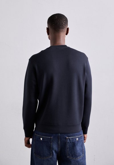 Juodas megztinis su apykakle, raibotais rankogaliais ir apačia. Suderintas su džinsais, turinčiais dvi galines kišenes ir matomu siuvimu.