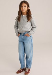 Siva žametna jopica z gumbnim zapiranjem, v kombinaciji s svetlo modrimi sproščenimi jeans hlačami. Rjave in rdeče vezene superge dodajo kontrast.