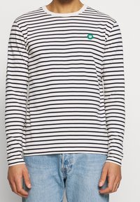 Långärmad t-shirt i vitt med marinblå ränder, med rund halsringning och en grön broderad logotyp på bröstet.