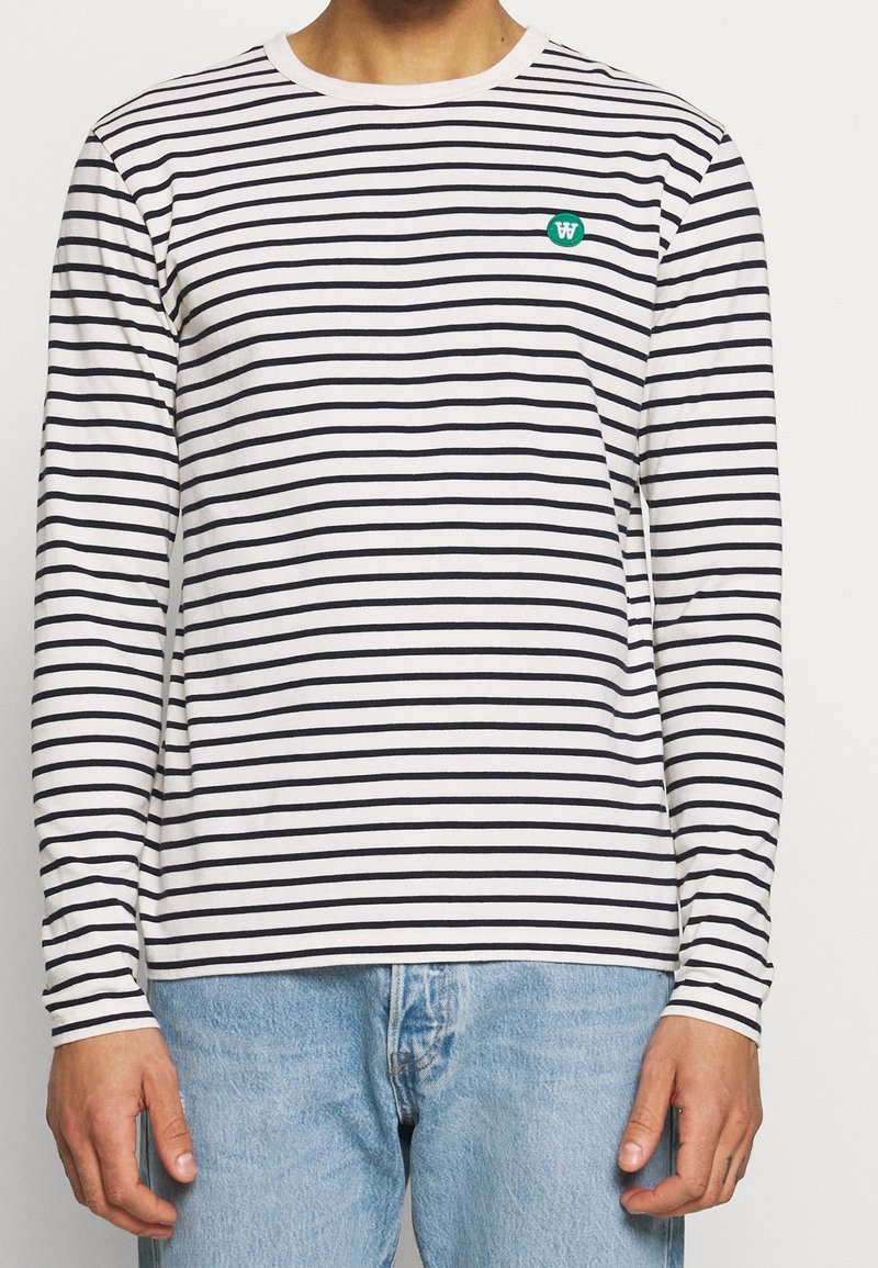 Långärmad t-shirt i vitt med marinblå ränder, med rund halsringning och en grön broderad logotyp på bröstet.