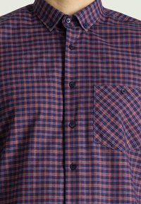 Camicia a quadri con motivi blu, rossi e bianchi. Presenta un colletto button-down e una tasca sul petto sinistro con un motivo a quadri diagonali.