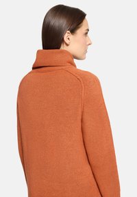 Femme aux cheveux bruns raides portant un pull orange en maille à col montant et manches longues, vue de profil sur un fond uni.