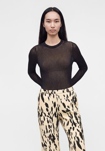Femme aux longs cheveux noirs porte un haut noir transparent à manches longues et un pantalon crème avec des taches abstraites noires, debout devant un fond uni.