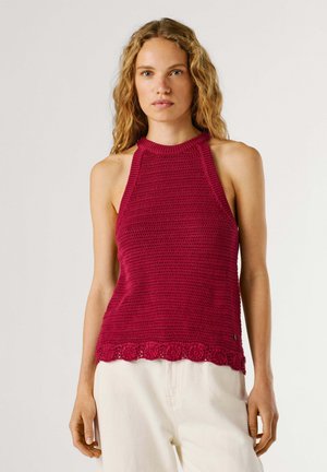 Jeune femme aux cheveux blonds ondulés portant un haut au crochet rouge sans manches à col halter et un pantalon beige clair, debout devant un fond uni.