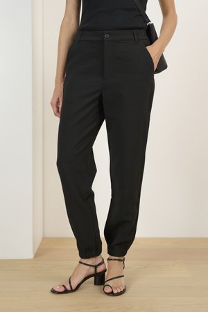 Black tapered trousers with elastic cuffs, side pockets, and button closure. Paired with black strappy heels, set against a light background.