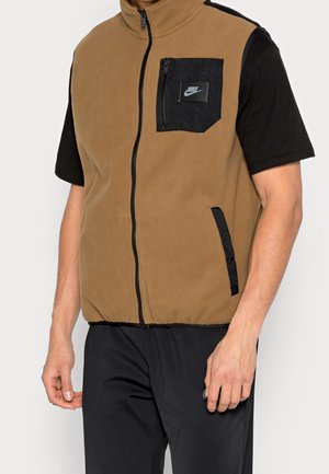 Hombre vistiendo un chaleco de forro polar marrón con bolsillo en el pecho con cremallera negra y bolsillos laterales, sobre una camisa de manga corta negra y pantalones negros.