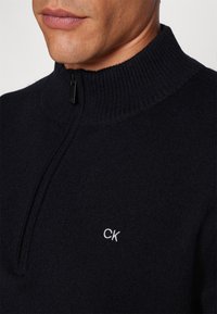 Sweater zippé marine avec col côtelé, fabriqué en matériau doux. Présente un logo blanc « CK » discret sur la poitrine. Design à coupe standard.