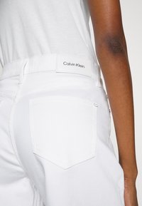 Primer plano de una persona que lleva vaqueros blancos de Calvin Klein y una camiseta blanca, mostrando el bolsillo trasero y la etiqueta de la marca en la cinturilla.