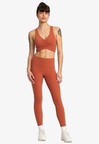Rustfärgad sportbh med rynkdetaljer och matchande högmidjade leggings, tillverkade av mjuk, elastisk tyg.