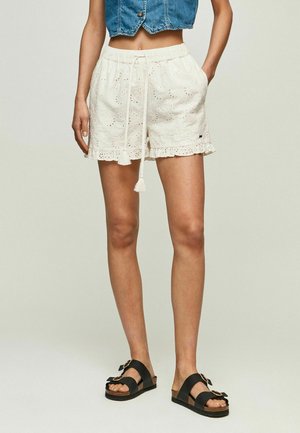 Torso femminile e gambe che indossano pantaloncini bianchi in sangallo con coulisse, gilet corto di jeans e sandali neri con doppia fascia e fibbie.