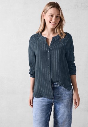 Camicia button-up a maniche lunghe di colore blu scuro con strisce verticali azzurro chiaro, caratterizzata da un colletto, una vestibilità comoda e maniche arrotolate. Indossata con jeans di denim chiaro.