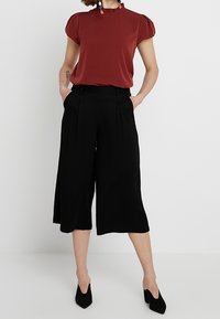 Blouse bordeaux à manches courtes avec un col froncé, associée à un pantalon culotte noir à jambes larges. Le mannequin porte des escarpins noirs à bout pointu, les mains dans les poches.