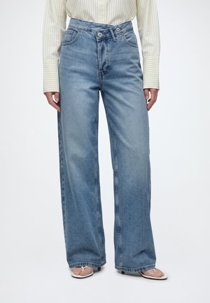 Personne portant un jean bleu taille haute à jambes larges avec une fermeture à double bouton, associé à une chemise rayée crème et des sandales blanches ouvertes à l'avant.