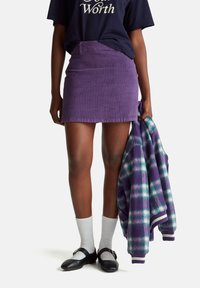 Mini falda de pana morada con un diseño ajustado, combinada con una camiseta oscura, calcetines blancos y zapatos negros, sosteniendo una chaqueta de franela.