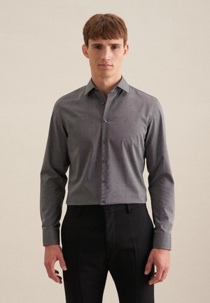 Jeune homme portant une chemise grise cintrée à manches longues rentrée dans un pantalon de costume noir sur un fond uni.