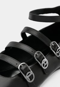 Zapato de cuero negro con tres tiras estrechas y hebillas plateadas, que presenta una textura suave y un diseño de punta redondeada.