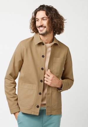 Beige jasje met een geknoopte voorkant, twee grote zakken en een kraag. Draagt over een lichtbeige trui met een gestructureerd patroon.