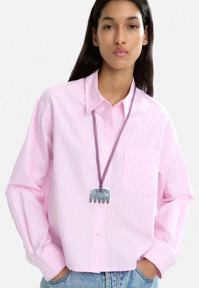 Jasnoróżowa koszula w paski z wyostrzonym kołnierzykiem, długimi rękawami i kieszenią na piersi. Prezentowany jest naszyjnik w kształcie srebrnego słonia.