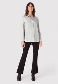 Eine Frau steht, trägt ein hellgraues Langarmhemd mit Knöpfen, schwarze Schlaghosen und schwarze Stiefeletten vor einem schlichten Hintergrund.