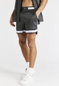 Shorts à motif gris avec des bandes blanches sur les côtés, une taille élastique et un bouton décoratif. Portés avec des baskets blanches et des chaussettes.