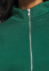 Zelená mikina na zip s vysokým límcem, s stříbrným zipem a texturovanou tkaninou, vhodná pro volnočasové nošení.