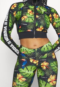 Urheilullinen lyhyt vetoketjullinen takki ja legginsit mustana, joissa on elävät trooppiset kukka- ja lehtikuosiset. Takissa on valkoista brändäystä hihoissa.
