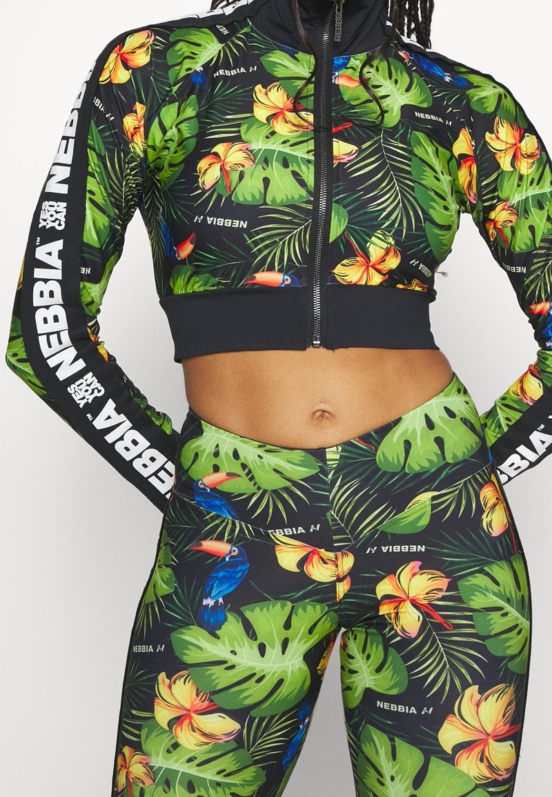 Urheilullinen lyhyt vetoketjullinen takki ja legginsit mustana, joissa on elävät trooppiset kukka- ja lehtikuosiset. Takissa on valkoista brändäystä hihoissa.