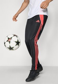 Black athletic pants featuring three vertical red stripes, made of smooth fabric. Worn with black soccer shoes; a soccer ball is nearby.