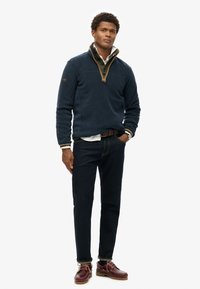 Marine fleece pullover med kvart-zipe, der har krave, parret med mørke denimjeans og brune bådsko.