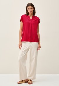 Blusa rossa a maniche corte con scollo a V e vestibilità rilassata, abbinata a pantaloni larghi crema e sandali marroni con cinturini su uno sfondo chiaro.