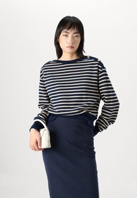 Pull rayé bleu marine et beige avec col rond, présentant des accents de boutons sur les épaules, assorti d'une jupe bleu marine ajustée et d'un petit sac blanc.