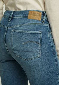 Blå denimjeans med en figurnära design, med en brun G-Star-logotyp, bakfickor och gula sömnadsdetaljer.