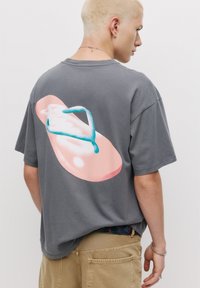 Grå t-shirt med et stort, farverigt grafik af en flip-flop trykt på ryggen. Korte ærmer, afslappet pasform, lavet af bomulds materiale.