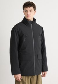 Giacca nera con colletto alto, zip frontale, due tasche al petto con zip e due tasche frontali con pattina. Tessuto liscio senza motivi visibili.