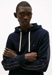 Marineblauwe hoodie van katoen, met crèmekleurige trekkoorden, rode en witte accenten langs de naden en een klein logo op de borst.