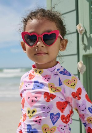 Kleines Kind trägt rote, herzförmige Sonnenbrille und ein buntes Hemd mit verschiedenen lächelnden Herzmustern, steht am Strand neben der Rettungsschwimmerhütte.