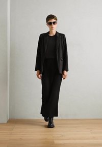 Černý přizpůsobený blazer přes černý top a kalhoty. Modelka nosí černé sluneční brýle a boty. Hladká textura, elegantní design. Světlá dřevěná podlaha.