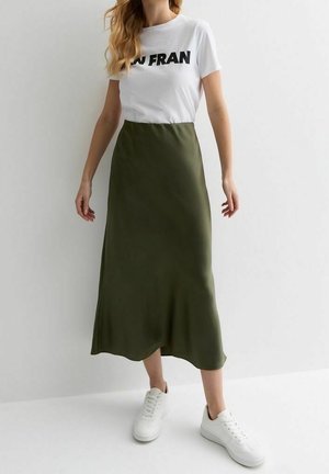 Mujer con camiseta blanca con estampado, falda midi verde oliva y zapatillas blancas, de pie contra un fondo claro y simple.