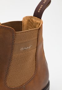 Nahaufnahme eines braunen Lederstiefels mit strukturiertem elastischen Seitenpaneel und einem Stoffziehhilfe, die Markendetails zeigt.