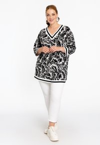 Zwart-witte bloemenpatroon tuniek met een V-hals, driekwart mouwen en contrasterende afwerking, gecombineerd met witte broek en schoenen.