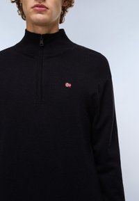Jeune personne portant un pull noir à demi-fermeture éclair avec un petit emblème de drapeau rouge, blanc et bleu sur la poitrine gauche sur un fond uni.