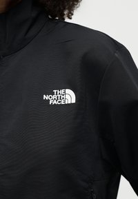 Άτομο που φορά μαύρο μπουφάν με λευκό κέντημα του λογότυπου The North Face στο αριστερό στήθος.