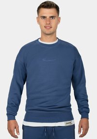 Felpa blu in un tessuto morbido, con polsini e orlo a coste, logo ricamato sul petto e design a strati con una camicia bianca sotto.