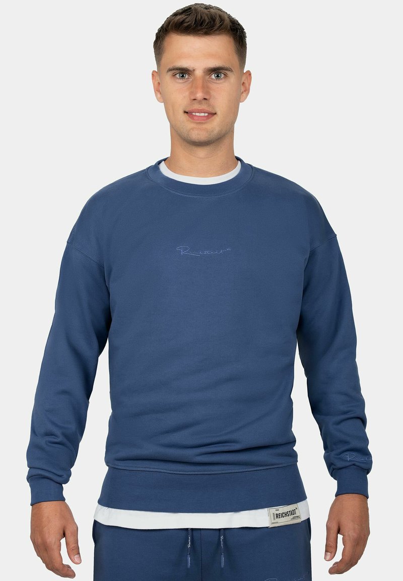 Felpa blu in un tessuto morbido, con polsini e orlo a coste, logo ricamato sul petto e design a strati con una camicia bianca sotto.