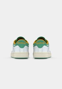 Biele kožené tenisky so zeleným a žltým pätovým dodatkom, štruktúrovanou podrážkou a logom "Reebok" v oranžovej farbe na zadnej strane.