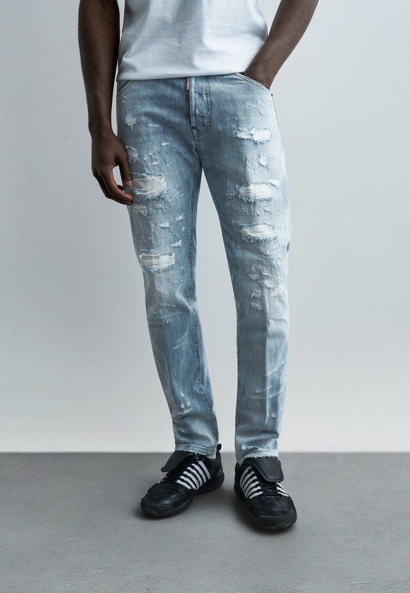 Homme portant un jean bleu clair délavé et des baskets noires avec des lacets blancs, debout devant un fond gris uni.